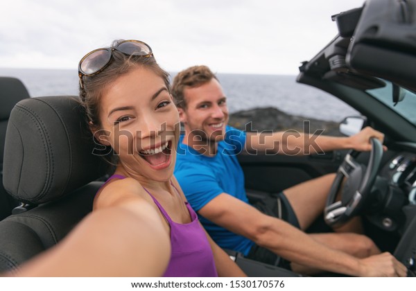 Selfie car travel couple having fun on\
summer road trip vacation driving to holidays. Asian woman and man\
excited taking self-portrait photo with mobile\
phone.