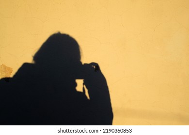 Selfie Of A Female Photographer In The Form Of A Human Shadow With A Camera On A Yellow Old Shabby Wall Background. Copy Space
