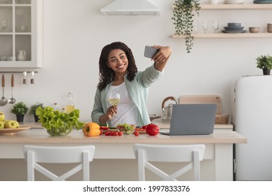 Selfie Of Food Blogger, Modern Cooking, Online Chat And Social Distance. Happy Young African American Female With Laptop, Takes Photo Or Video, Holds Glass Of Wine In Kitchen Interior With Vegetables