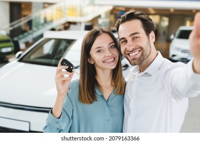 Selfie On Smart Phone Of Happy Young Family Holding Car Keys After Buying New Automobile At Dealer Shop Store. Heterosexual Husband And Wife Purchasing Expensive Auto