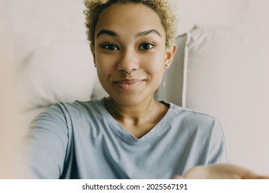 Selfie on white home background of dark skinned trendy looking blonde teenager with nose and ears jewelry, making happy, excited and surprised face expression, wearing blue long-sleeved shirt