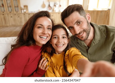 Selfie With Parents. Happy kid daughter making selfie photo with daddy and mommy, smiling to camera posing together for portrait at home interior. Family having fun taking photo of themselves - Powered by Shutterstock