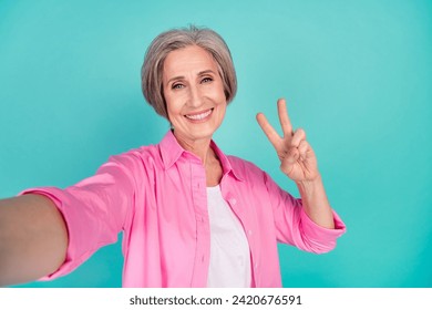 Selfie photo of cheerful happy grandmother makes her own blog demonstrate v sign greetings isolated on aquamarine color background