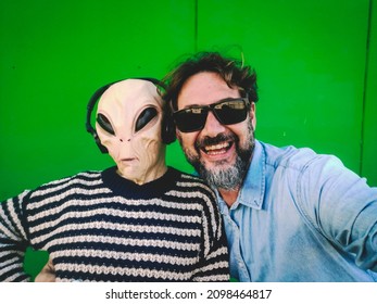 Selfie Portrait With Cheerful Happy Adult Man Smiling And Serious Funny Alien Mask With Headphones. Green Wall Background. Concept Of Ufo Estraterrestrial And Human People Together