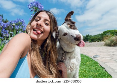Selfie portrait of young Colombian Latina woman, with her border collie dog, in the park sticking out her tongue and looking at the camera, with the sky and trees in the background, phone perspective. - Shutterstock ID 2254338353