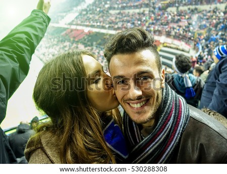 Selfie portrait of young couple watching sport soccer match in football italian stadium - Young people having fun together - Love for sport concept - Focus on man - Warm filter with stark arena lights