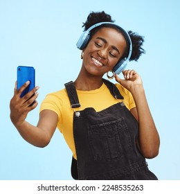 Selfie, smartphone and black woman with headphones, social media and connection on blue studio background. African American female influencer, girl and content creator with smile and online streaming - Shutterstock ID 2248535263