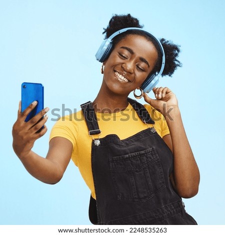 Selfie, smartphone and black woman with headphones, social media and connection on blue studio background. African American female influencer, girl and content creator with smile and online streaming
