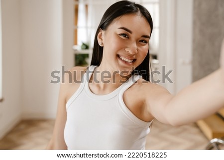 Selfie of young beautiful smiling happy asian woman in white sleeveless t-shirt looking at camera, while standing at home