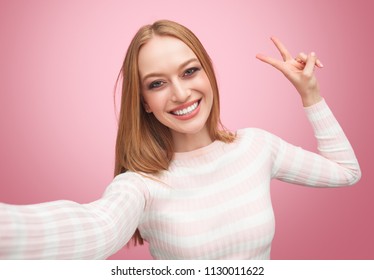 Selfie Of Young Cute Female Smiling Showing Peace Sigh With Fingers And Looking At Camera In Studio With Pink Background