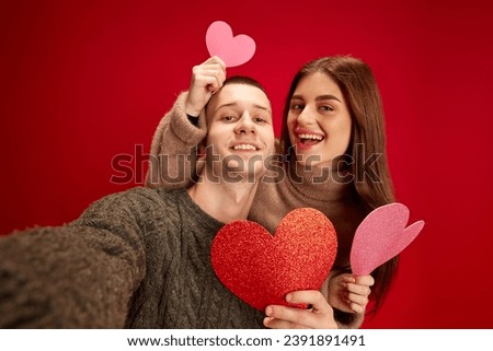 Selfie. Young happy smiling man and woman, couple, boyfriend and girlfriend posing with paper hearts against red studio background. Concept of love, relationship, Valentine's Day, emotions, lifestyle