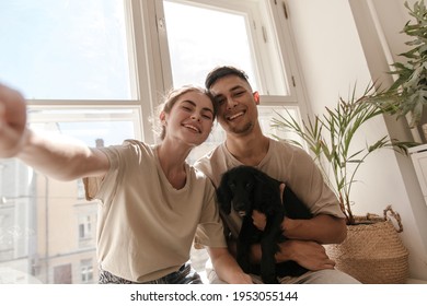 Selfie-portrait of young couple with black dog in light room against window background. Attractive girl wearing beige clothes smiling and posing near boyfriend, holding little puppy - Powered by Shutterstock