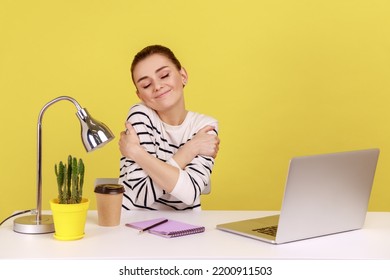 Selfish Happy Woman Office Manager Embracing Herself With Pleasure Expression, Sitting At Workplace, Feeling Proud And Satisfied. Indoor Studio Studio Shot Isolated On Yellow Background.