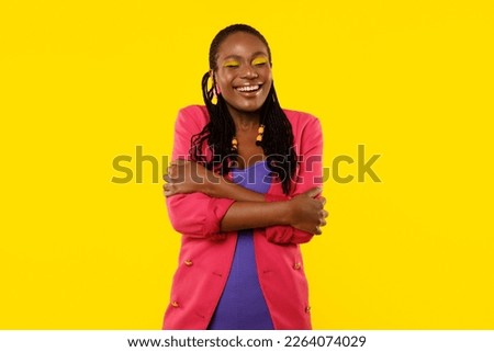 Self-Love. Happy African American Woman Hugging Herself Posing With Eyes Closed Standing Over Yellow Studio Background, Wearing Bright Clothes. Accept Your True Self Concept