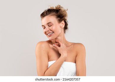 Self-Pampering Concept. Middle Aged Woman Wrapped In Bath Towel Touching Her Skin While Standing Over Light Gray Background, Attractive Mature Lady Enjoying Beauty Treatments, Copy Space