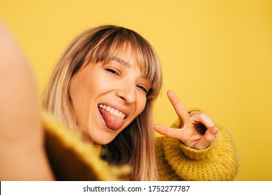 Self-portrait of young blonde woman posing on camera and taking selfie. Holding two fingers up in peace symbol. Girl in yellow sweater showing tongue. Isolated over background - Powered by Shutterstock