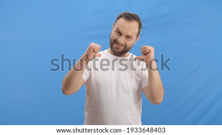A self-righteous happy young man in front of the blue background, pointing and showing himself with his thumbs. Self-pointing concept in front of a blue background.