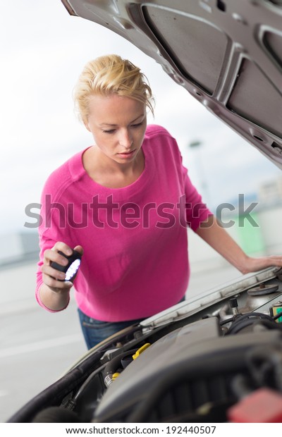 Self-sufficient confident modern\
young woman inspecting broken car engine with a flashlight in her\
hand. 