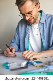 A self  taught seamster and glasses is working at the table  A DIY designer draws sketch for new costume project  A smiling man in blue shirt makes sketch for sewing clothes as hobby 