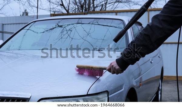 self-washing\
the car with a brush and the pressure of water from a pistol in the\
yard in the evening, a man manually cleaning a gray car on the\
street at dusk using a high pressure\
washer