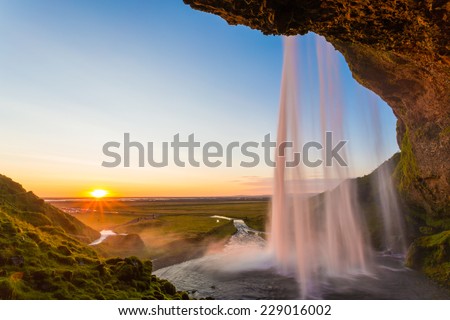 Seljalandsfoss waterfall at sunset, view from behind the fall, sunburst and sunstar, iceland. clear blue sky
