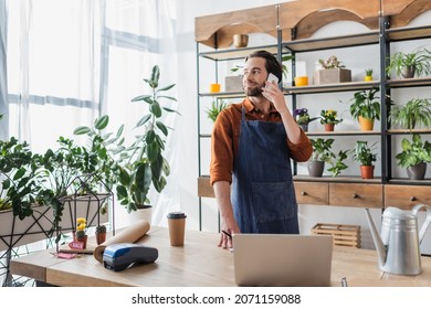 Seller in apron talking on smartphone near coffee and laptop in flower shop - Powered by Shutterstock