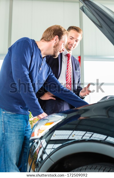 Seller or car salesman and\
client or customer in car dealership presenting the engine\
performance of new and used cars in the showroom, the men looking\
under the hood