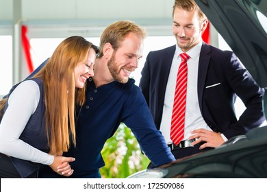 Seller Or Car Salesman And Clients Or Customers In Car Dealership Presenting The Engine Performance Of New And Used Cars In The Showroom, The Men Looking Under The Hood