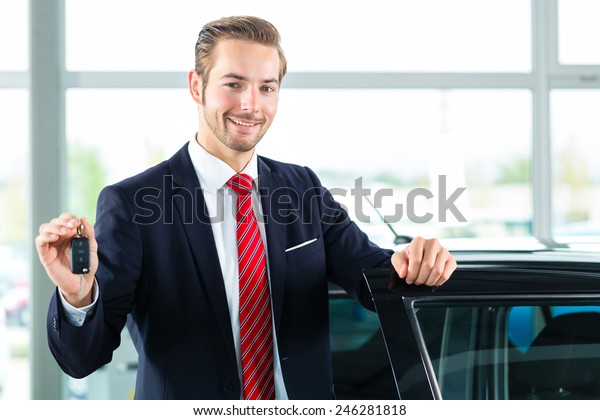 Seller or car salesman in car\
dealership with key presenting his new and used cars in the\
showroom