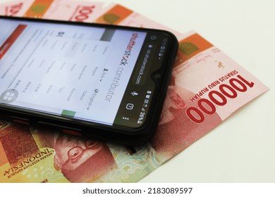 Semarang, Indonesia - July 27, 2022: An Android phone screen image displays the "Shutterstock Contributor" website with a background visualization of the 100,000 Indonesian rupiah currency.