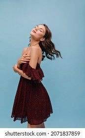 Semi sideways pretty young woman stands on blue background with her head tilted back with closed eyes. Light-skinned lady with bright lipstick is dressed in short summer dress with bare shoulders.