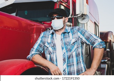 Semi Truck Professional Driver On The Job In Casual Clothing Wears Safety Medical Face Mask. Confident Looking Trucker Stands Next To Red Big Rig Wearing Protection Sunglasses And Surgical Mask.