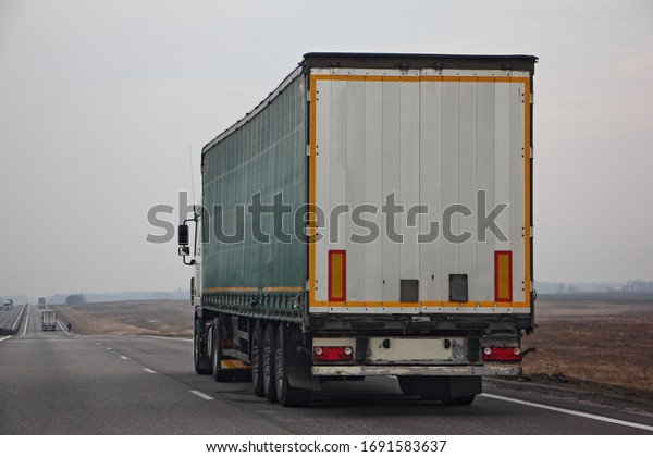 Semi truck van drive on dry suburban\
asphalt highway at spring day, rear-side view – international\
logistics, cargo transportation, trucking\
industry