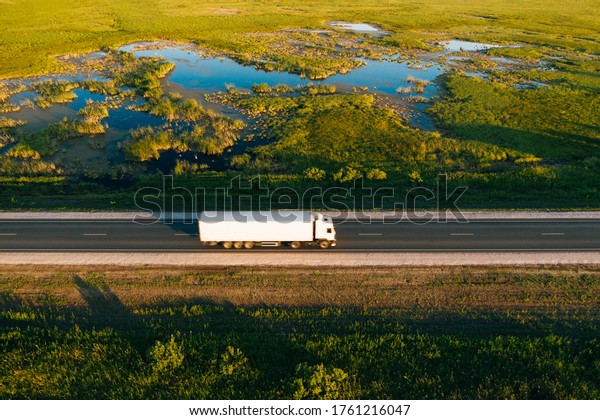 Semi\
truck with white freight trailer drive on highway in green grass\
countryside at sunrise, aerial top view. Car with motion blur\
effect. Road transportation and logistics\
background