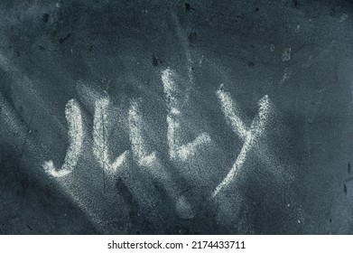 Semi-erased Word JULY On Black Chalkboard. Handwritten Word. Fuzzy Letters On A Black Surface. The Concept Of Alternating Months Of The Year. Calendar