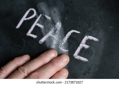 The Semi-erased Word PEACE On The Black Chalkboard. An Adult Man's Left Hand Removes The Handwritten Word With His Fingers. Fuzzy Letters On Black Surface.