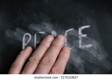 The Semi-erased Word PEACE On The Black Chalkboard. An Adult Man's Left Hand Removes The Handwritten Word With His Fingers. Fuzzy Letters On Black Surface.