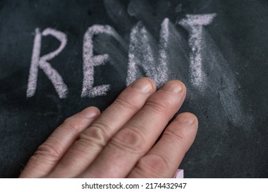 The Semi-erased Word RENT On The Black Chalkboard. An Adult Man's Left Hand Removes The Handwritten Word With His Fingers. Fuzzy Letters On Black Surface.