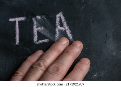 The Semi-erased Word TEA On The Black Chalkboard. An Adult Man's Left Hand Removes The Handwritten Word With His Fingers. Fuzzy Letters On Black Surface.