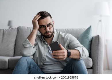 Sending Error, Bad News, Feeling Stress And Heartbreak From Relationships. Disappointed Sad Millennial Man In Glasses Sits On Floor Looks At Phone And Pressed Hand To Head In Living Room Interior