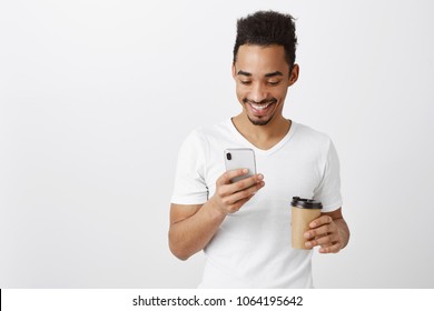 Sending Funny Meme To Girlfriend. Portrait Of Handsome African Coworker In Casual White Shirt, Holding Cup With Coffee And Smartphone, Typing Message Or Playing Game, Having Fun Over Gray Background