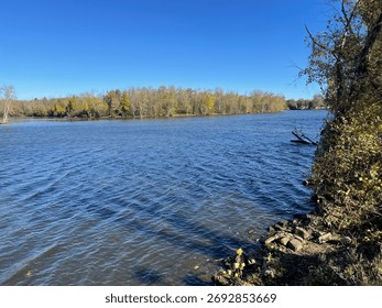 The Seneca River flows calmly under a clear blue sky, surrounded by trees showing early autumn colors in Baldwinsville, New York.