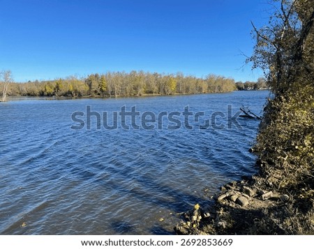 The Seneca River flows calmly under a clear blue sky, surrounded by trees showing early autumn colors in Baldwinsville, New York.