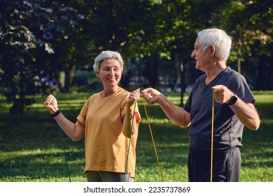 Senior 60 spouses wear sportswear doing exercises, training arms outdoor in summer park in the morning using resistance rubber bands. Healthy lifestyle, active retired life use modern appliance - Powered by Shutterstock