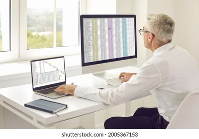Senior accountant man using laptop and desktop computer while working in modern office. Mature white male company employee and team project manager sitting at desk, looking at screen 