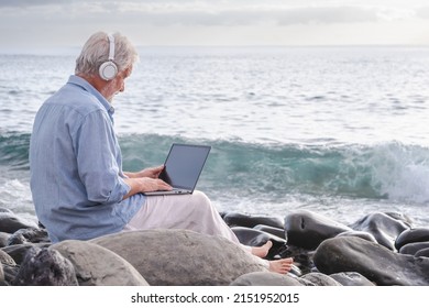 Senior Active Man Sitting On The Beach Using Laptop Wearing Headphones. Elderly Bearded Male Barefoot Enjoying Sunset At Sea, Horizon Over Water