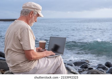 Senior Active Man Sitting On The Beach Using Laptop. Elderly Bearded Male Holding A Coffee Cup Enjoying Sunset At Sea, Horizon Over Water