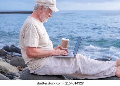 Senior Active Man Sitting On The Beach Barefoot Using Laptop. Elderly Bearded Male Holding A Coffee Cup Enjoying Sunset At Sea, Horizon Over Water