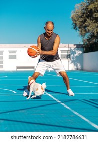 Senior adult athletic man holding a sport ball playing basketball with cute dog jack russel terrier on blue color playground outdoor at summer sunny day - Shutterstock ID 2182786631