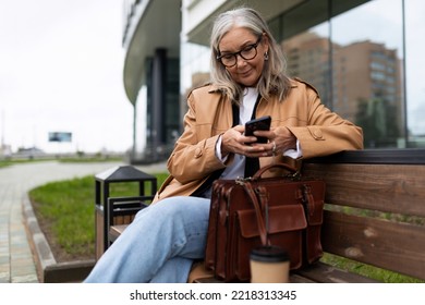 An Senior Adult Elderly Woman Sits In A Park On The Street And Reads A Message On A Mobile Phone, 50 Year Old European Woman